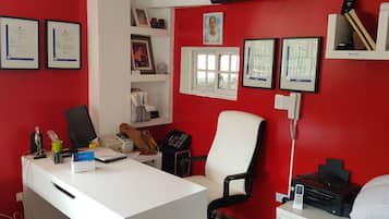 Una habitación con pared roja, escritorio blanco, dos sillas, certificados enmarcados y una ventana.