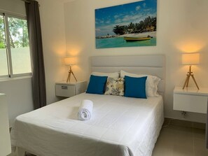 Una cama bien hecha con una toalla blanca enrollada, dos almohadas azules y un cabecero blanco. Hay dos mesitas de noche con lámparas y un cuadro de una playa sobre la cama.