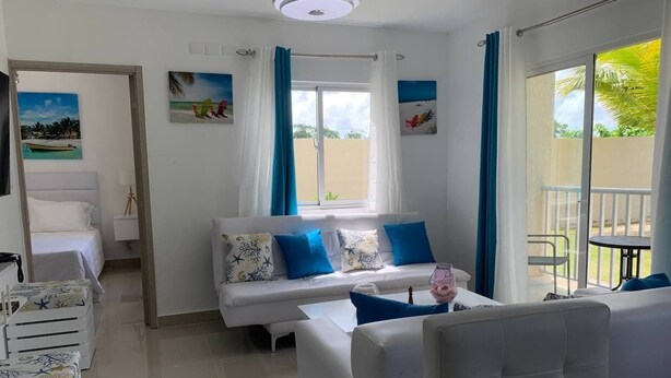 A modern living room with a white sofa set, a coffee table, and a dining area with a table and chairs. There is a balcony with a view of palm trees. - Conchita de Mar (Punta Cana)