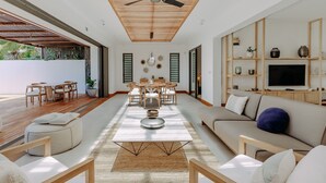 Un amplio salón con techo de madera, una mesa de comedor con sillas, un sofá y un televisor sobre una estantería de madera.
