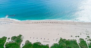 Une vue aérienne d’une plage aux eaux turquoise et limpides, avec un rivage sablonneux et des parasols disposés çà et là.