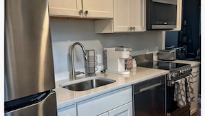 A modern kitchen with stainless steel appliances, white cabinetry, a microwave, and a coffee maker. - Roomy Themed 3BR Historic Salem, Near Boston, with Parking, Walk to Attractions. (Salem)