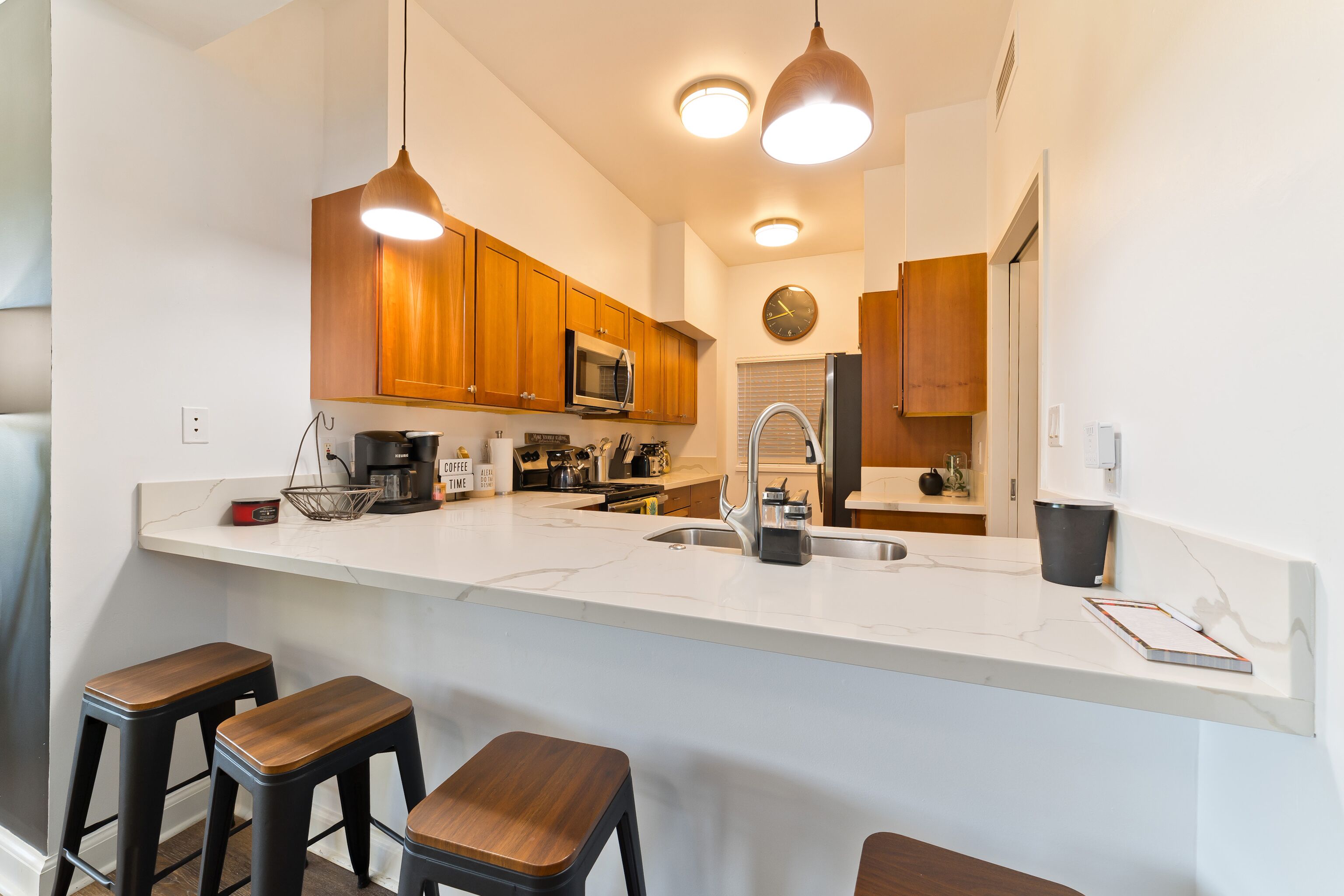 A modern kitchen with wooden cabinets, a white countertop, a built-in microwave, a stainless steel sink, and three wooden stools.