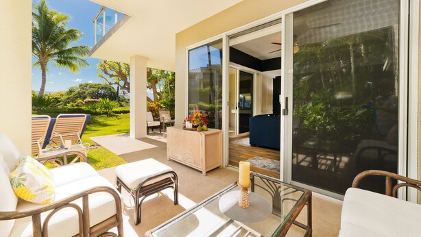 Un patio iluminado por el sol con mobiliario exterior, una mesa de vidrio y una puerta corrediza de cristal que da a un exuberante jardín.