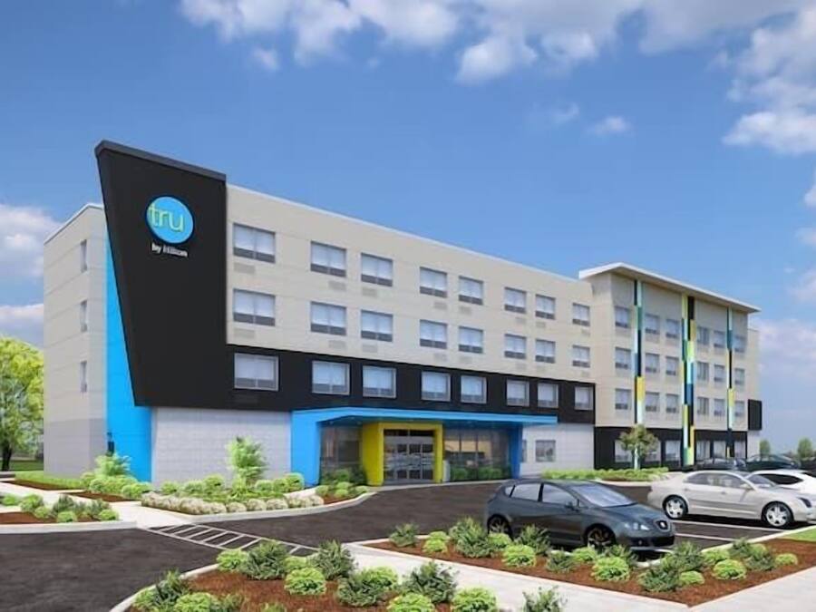 Un hôtel moderne, avec un logo bien visible sur le côté, plusieurs étages et un parking où sont garées plusieurs voitures.