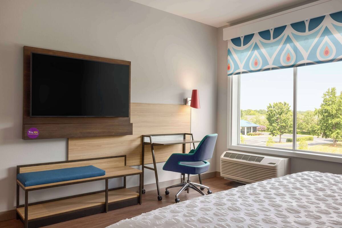 Une chambre d’hôtel dotée d’une grande télévision à écran plat, d’un bureau avec une chaise, d’un banc, d’un lit avec une couvre-lit à motifs et d’une fenêtre donnant sur un paysage verdoyant.