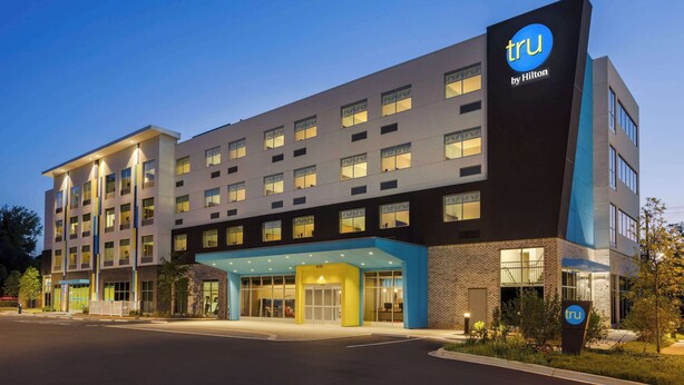 Un hôtel moderne, reconnaissable à son enseigne « tru by Hilton » bien visible, avec de nombreux étages et une entrée bien éclairée.