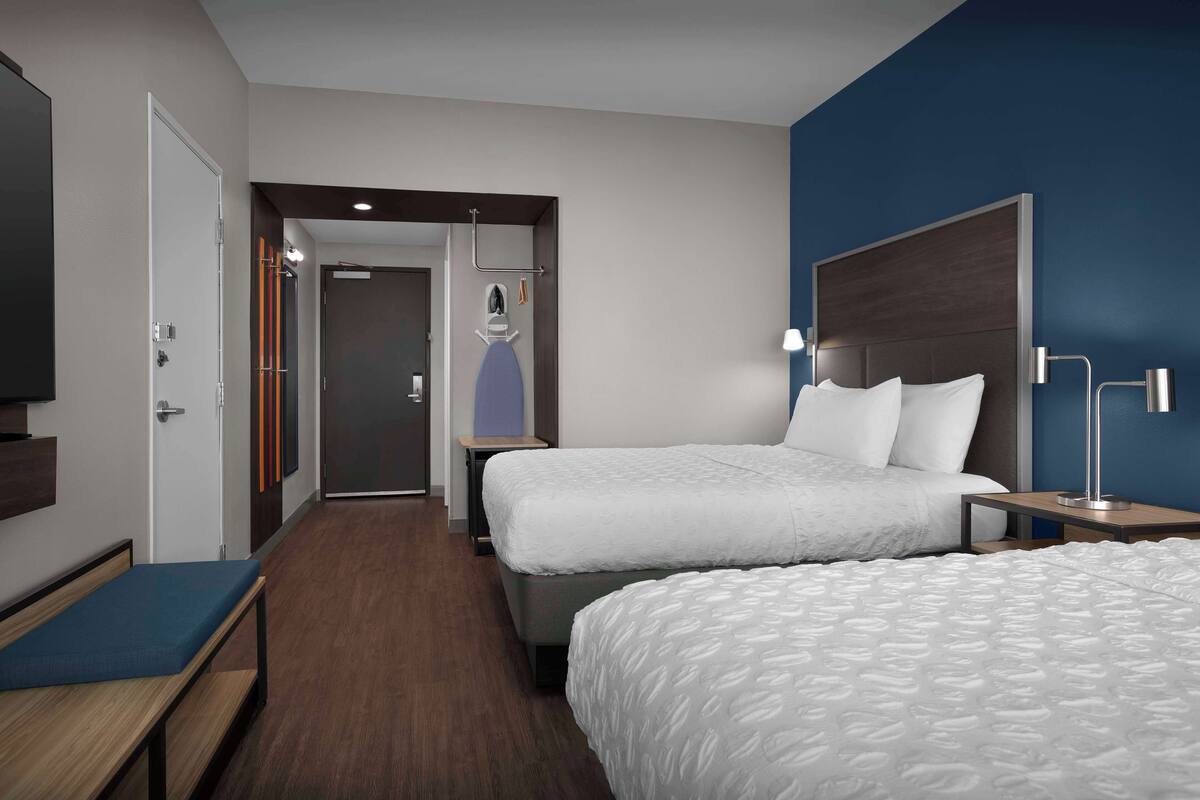 A hotel room with two beds, a bench, a TV, and a bathroom visible through an open door.