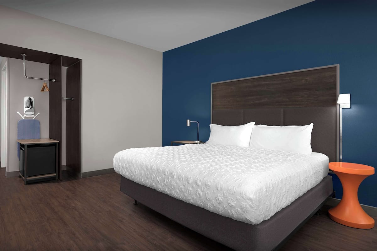 A modern hotel room with a large bed, a nightstand, a small table, and a wall-mounted lamp.