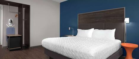A modern hotel room with a large bed, a nightstand, a small table, and a wall-mounted lamp.