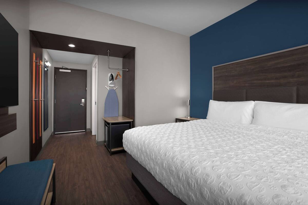 A hotel room with a large bed, a bedside table, a nightstand, a wall-mounted TV, and a door leading to another room.