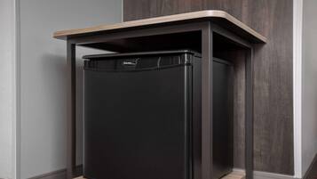 A black refrigerator with a wooden top shelf, placed in a corner against a gray wall and wooden panel.