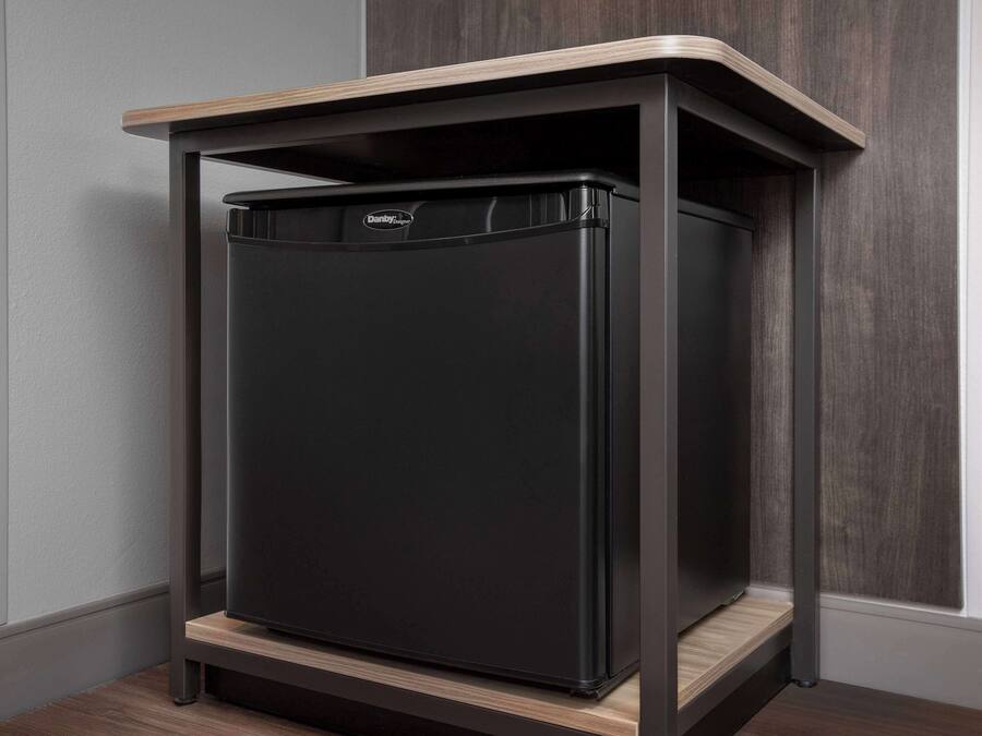 A black refrigerator with a wooden top shelf, placed in a corner against a gray wall and wooden panel.