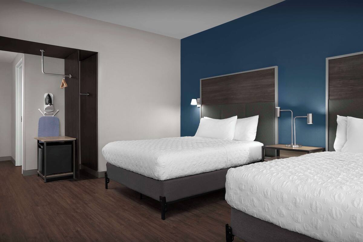 A hotel room with two beds, a wooden floor, a dark blue accent wall, and a small table with a lamp.