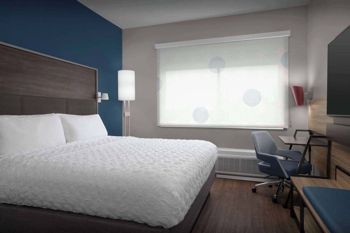 A hotel room with a large bed, a desk, a chair, a lamp, and a window with blinds.