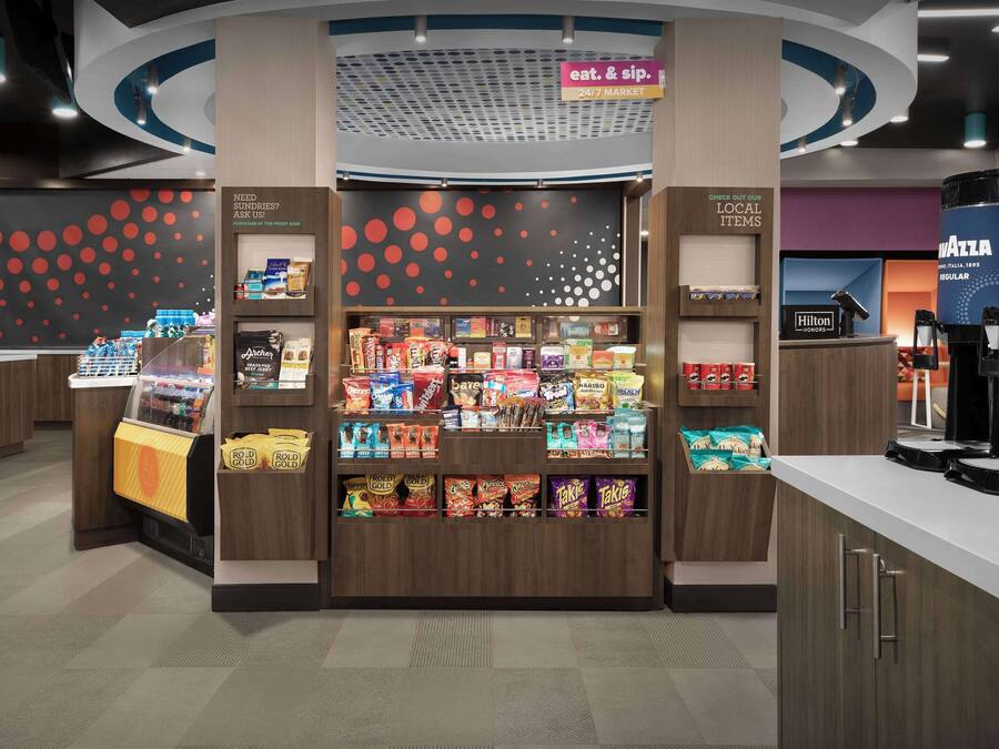 A snack kiosk with a variety of packaged foods and beverages, a 24/7 market sign, and a Hilton logo indicating nearby hotel services.