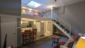 A compact living space with a kitchenette, a dining area with two chairs, a small table, a sofa, and a staircase with decorative railings. - Cozy Loft Hideaway in Robinson (Robinson)