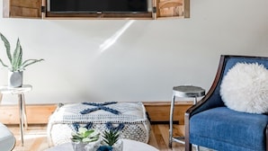 Una sala de estar con un televisor montado, un sillón azul, una mesa de centro redonda con objetos decorativos y una mesita auxiliar.