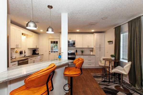 A modern kitchen with white cabinets, a marble countertop, and two orange bar stools.
