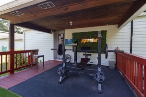 Ett utomhusgym med en bänkpress, ett knäböjsställ och vikter, placerat på en täckt terrass med ett rött räcke.