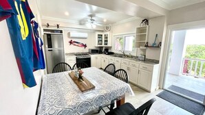 A kitchen with white cabinets, a microwave, and a dining table with chairs. There is a ceiling fan and a window with a view of greenery. - Utila Suite Sleeps 6 (Utila)