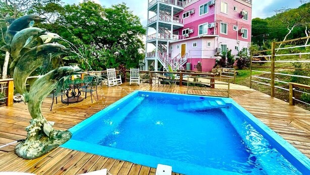 A swimming pool with a wooden deck, surrounded by a pink and white building with balconies. - Utila Suite Sleeps 6 (Utila)