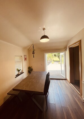 A dining area with a long table, chairs, a hanging light fixture, a window with blinds, and a view through a glass door. - River Cottage. Traditional Irish Cottage in Mullingar Ireland (Mullingar)
