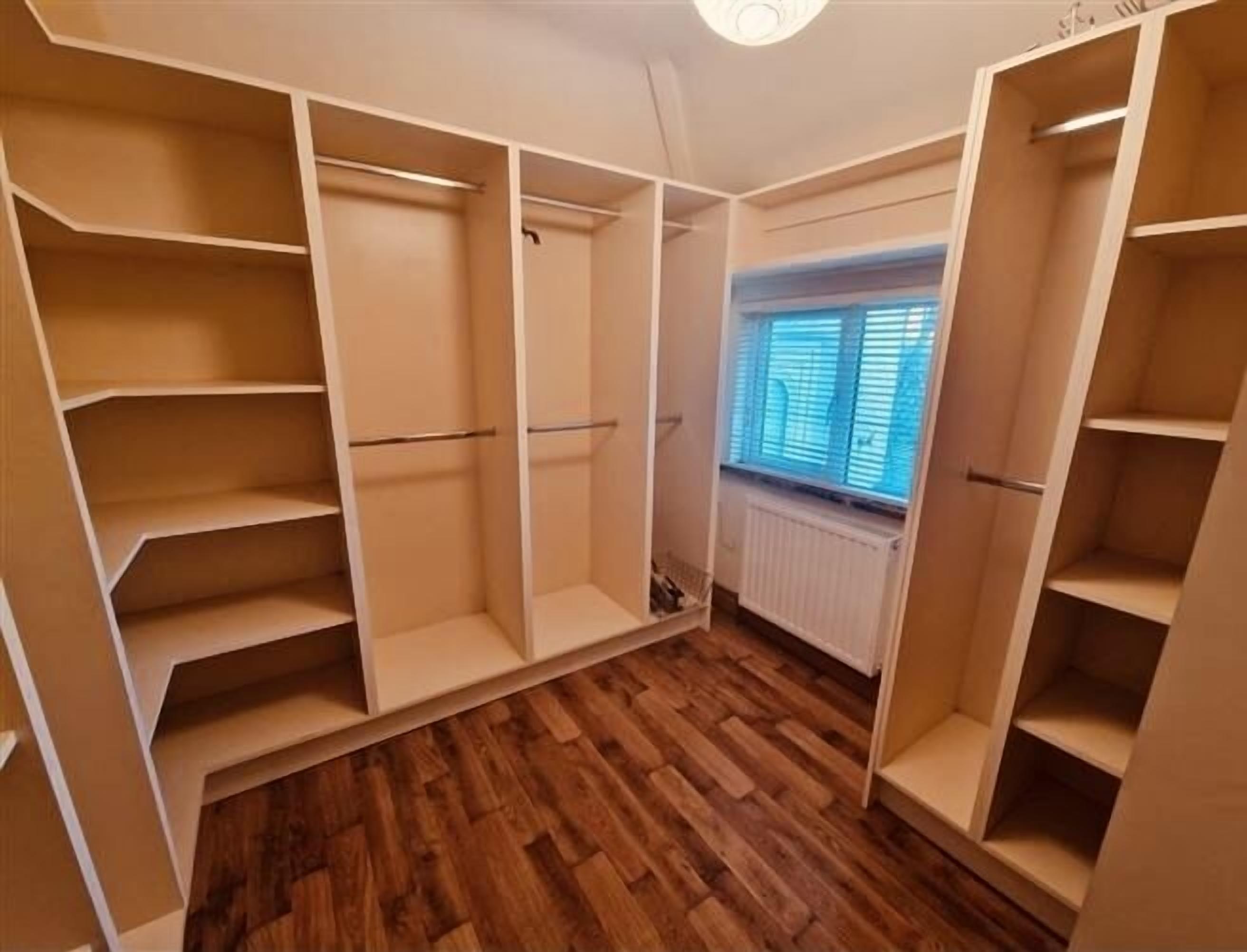A walk-in closet with multiple shelves and hanging spaces, wooden flooring, and a window with blinds.