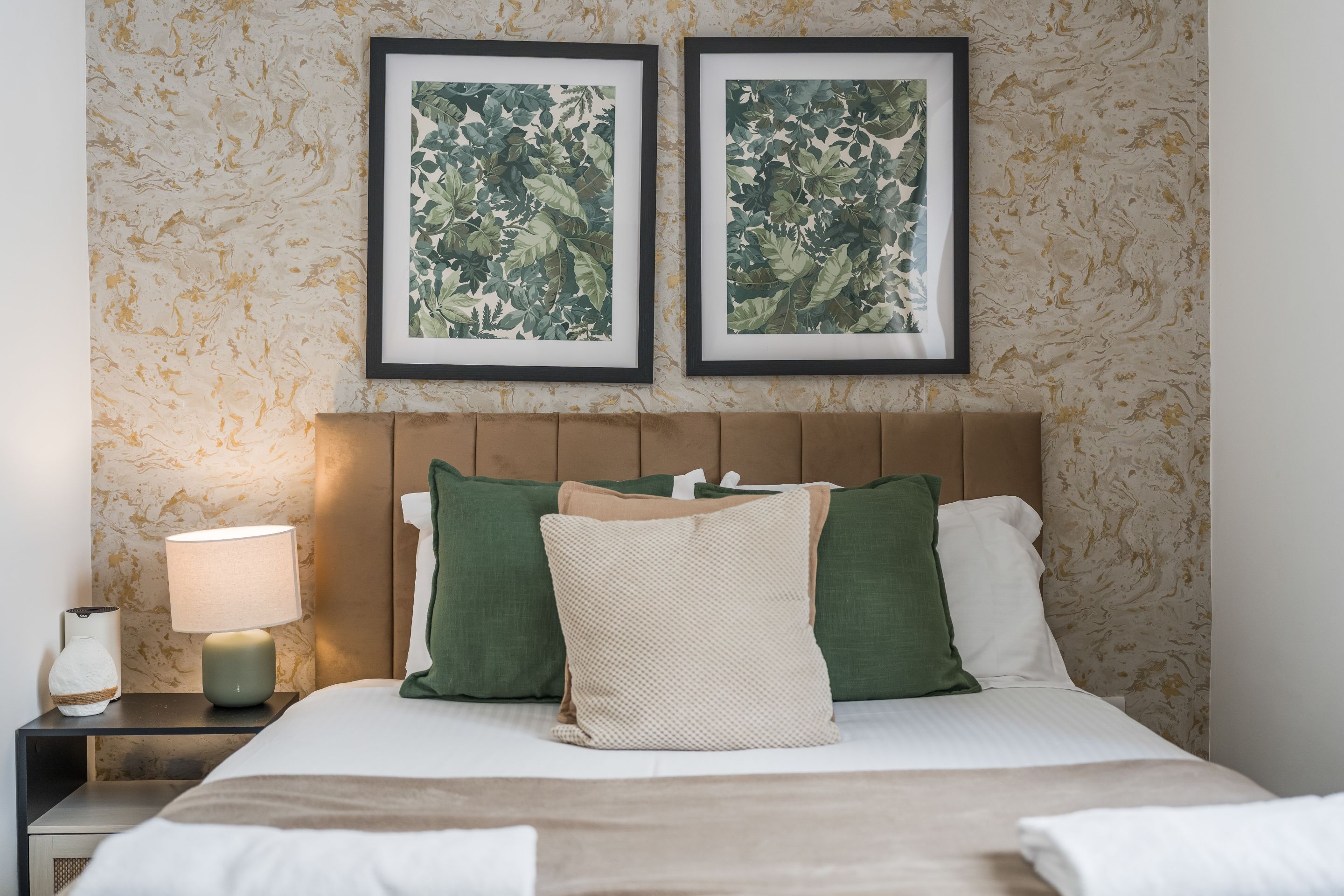A hotel room with a bed, two framed botanical prints on the wall, a nightstand with a lamp, and a bedside table with a vase.