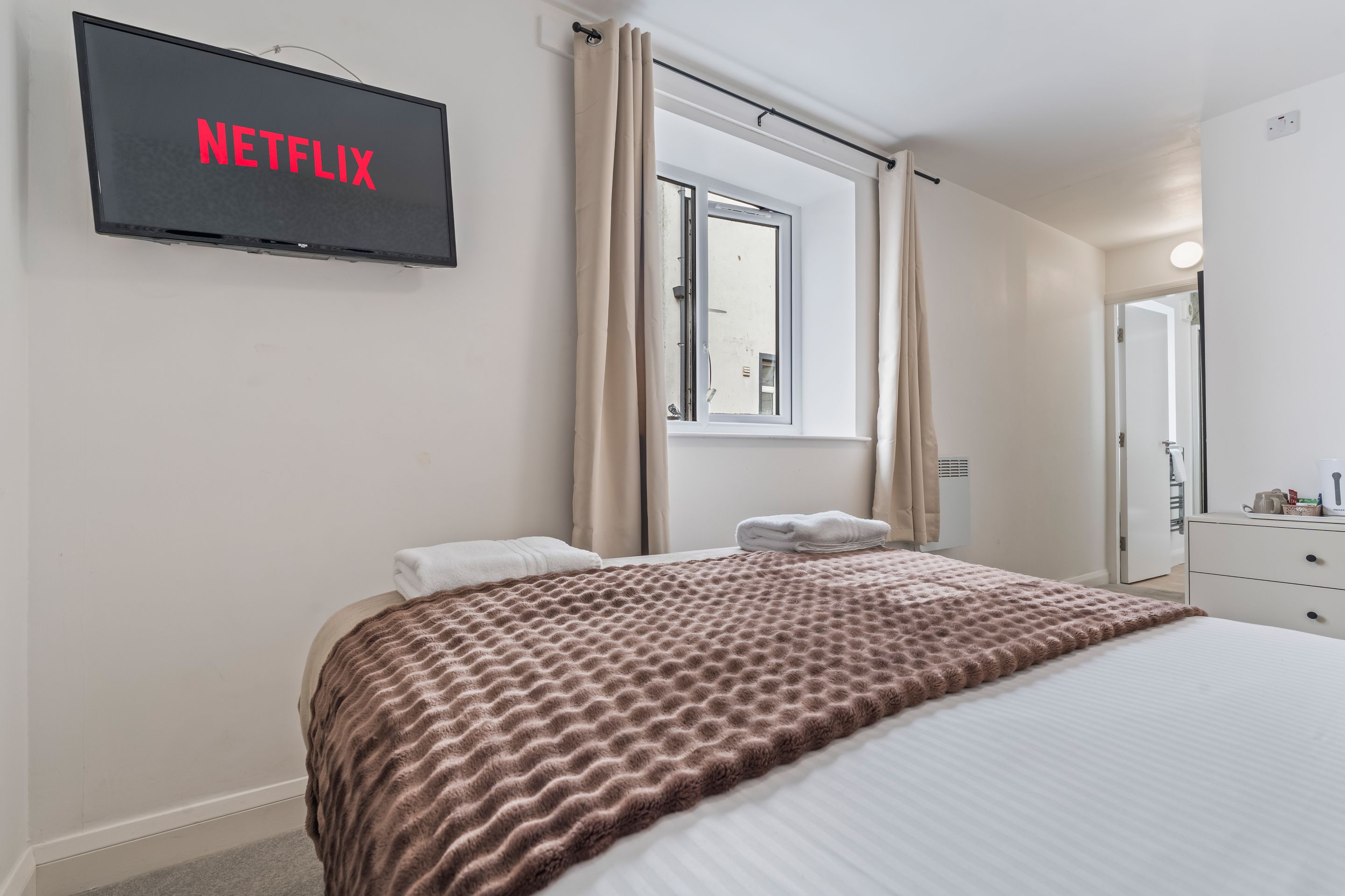 Una habitación de hotel con cama, televisión montada en la pared, ventana con cortinas, mesita de noche y espejo.