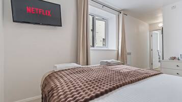 Una habitación de hotel con cama, televisión montada en la pared, ventana con cortinas, mesita de noche y espejo.