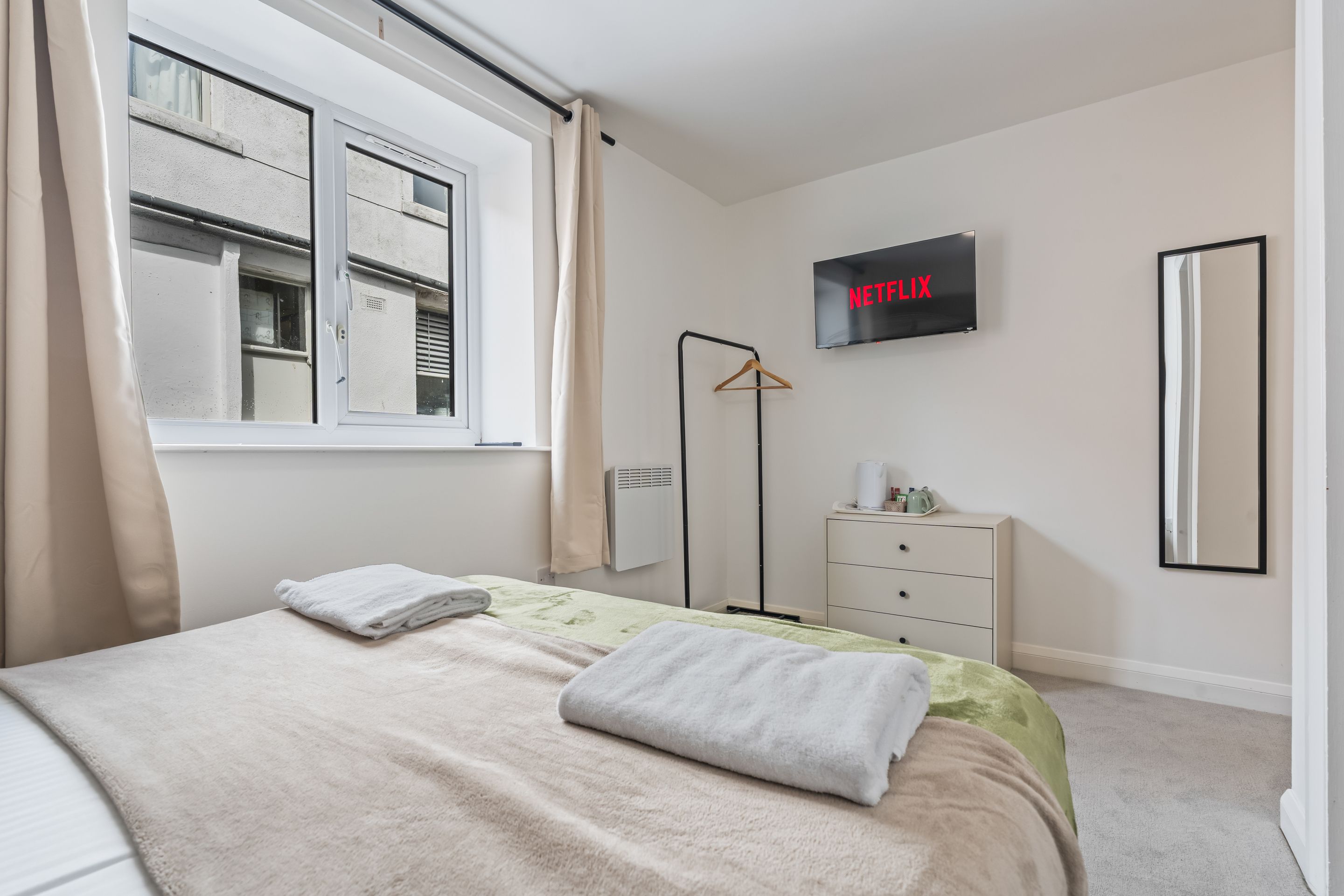 Un dormitorio con cama, televisión en la pared, cómoda y ventana con cortinas.