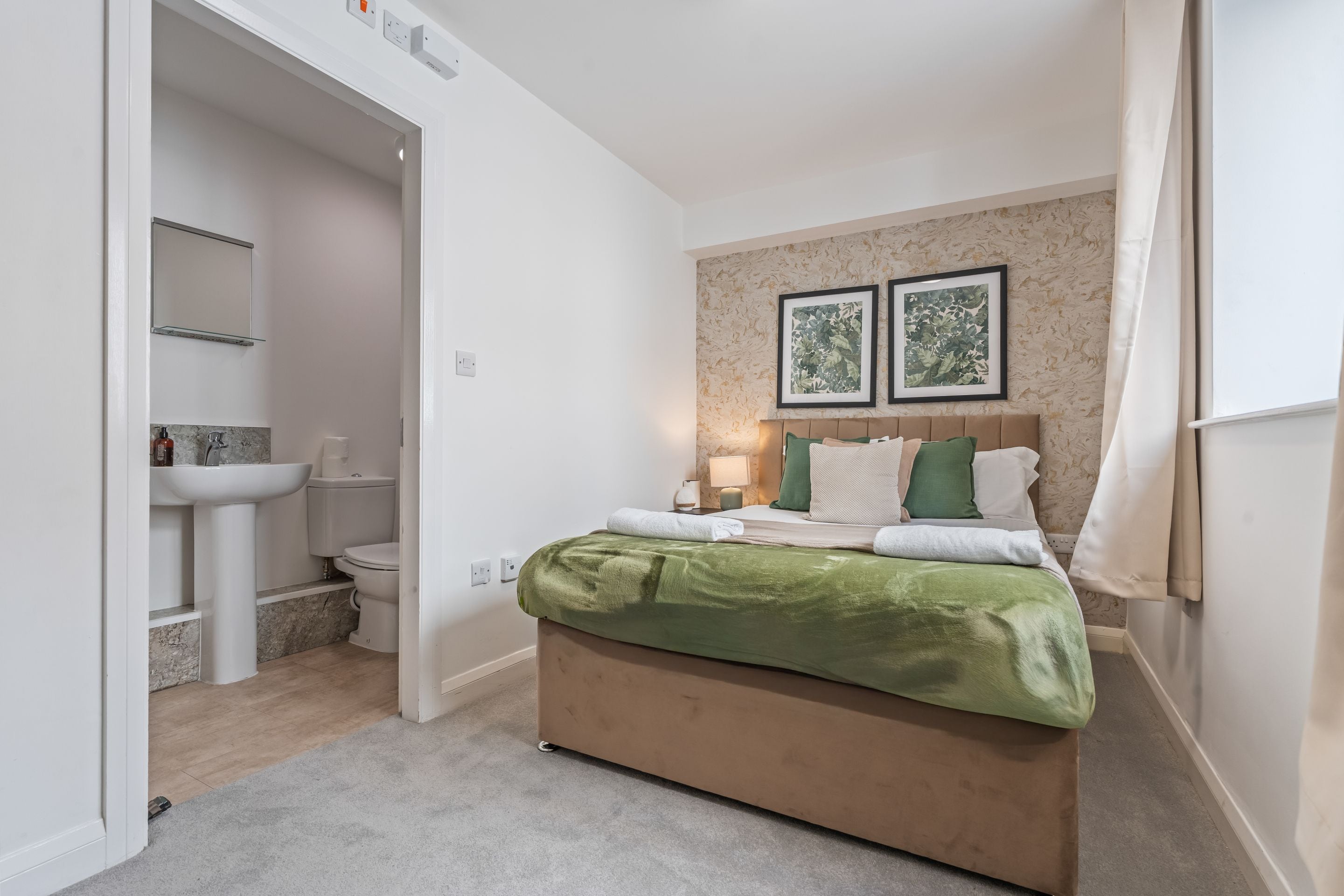 A bedroom with a bed, a nightstand, a lamp, a wall-mounted shelf, and a bathroom with a toilet and sink.
