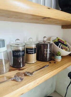 Un estante con recipientes etiquetados de café, azúcar moreno y artículos reutilizados, junto con una pequeña planta y una cuchara.
