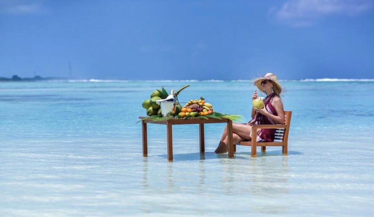A person seated at a wooden table with a tropical drink, surrounded by coconuts and fruits, on a beach with clear blue water.