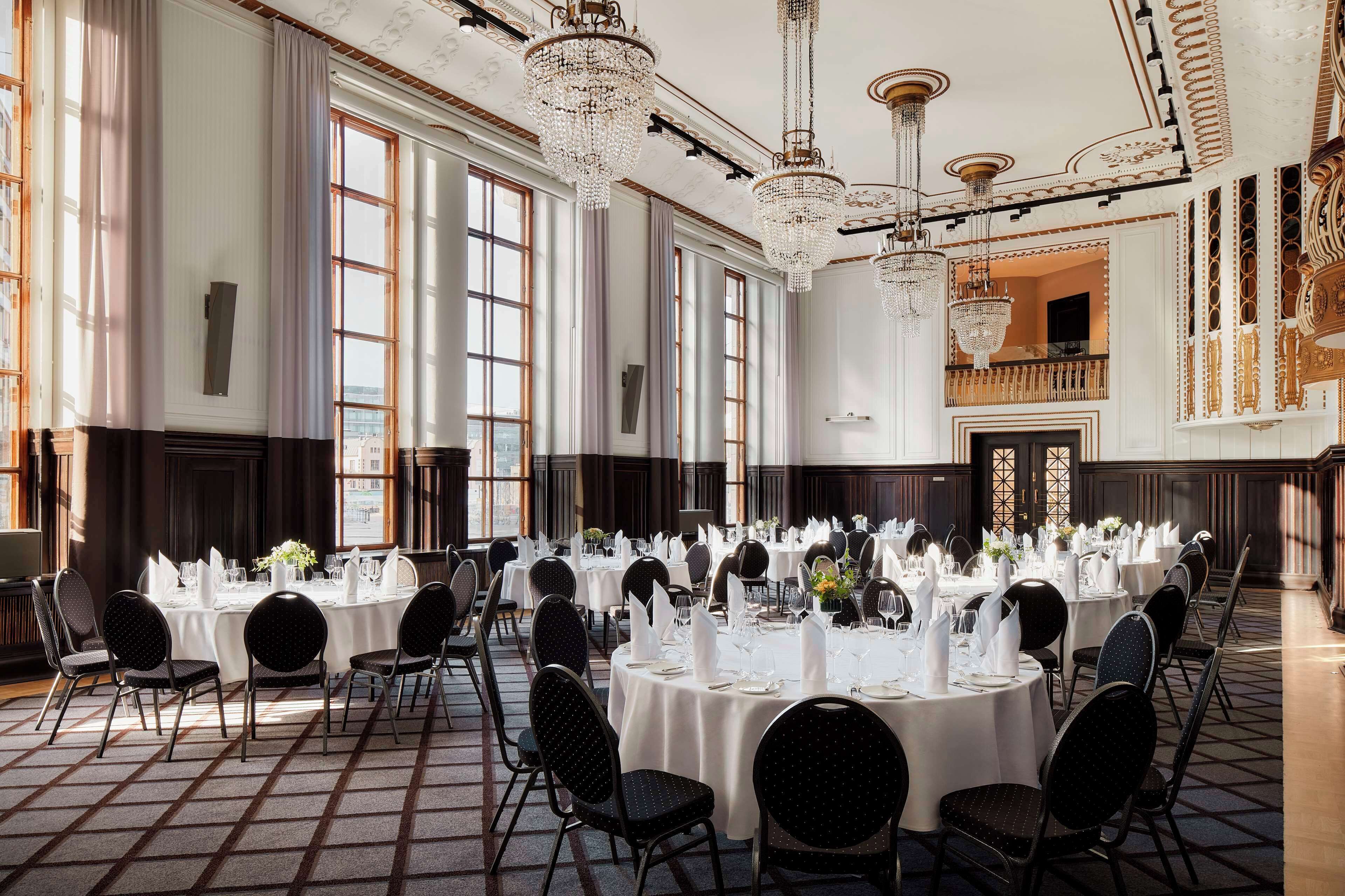 Een balzaal met ronde tafels gedekt voor een diner, grote ramen en versierde kroonluchters.