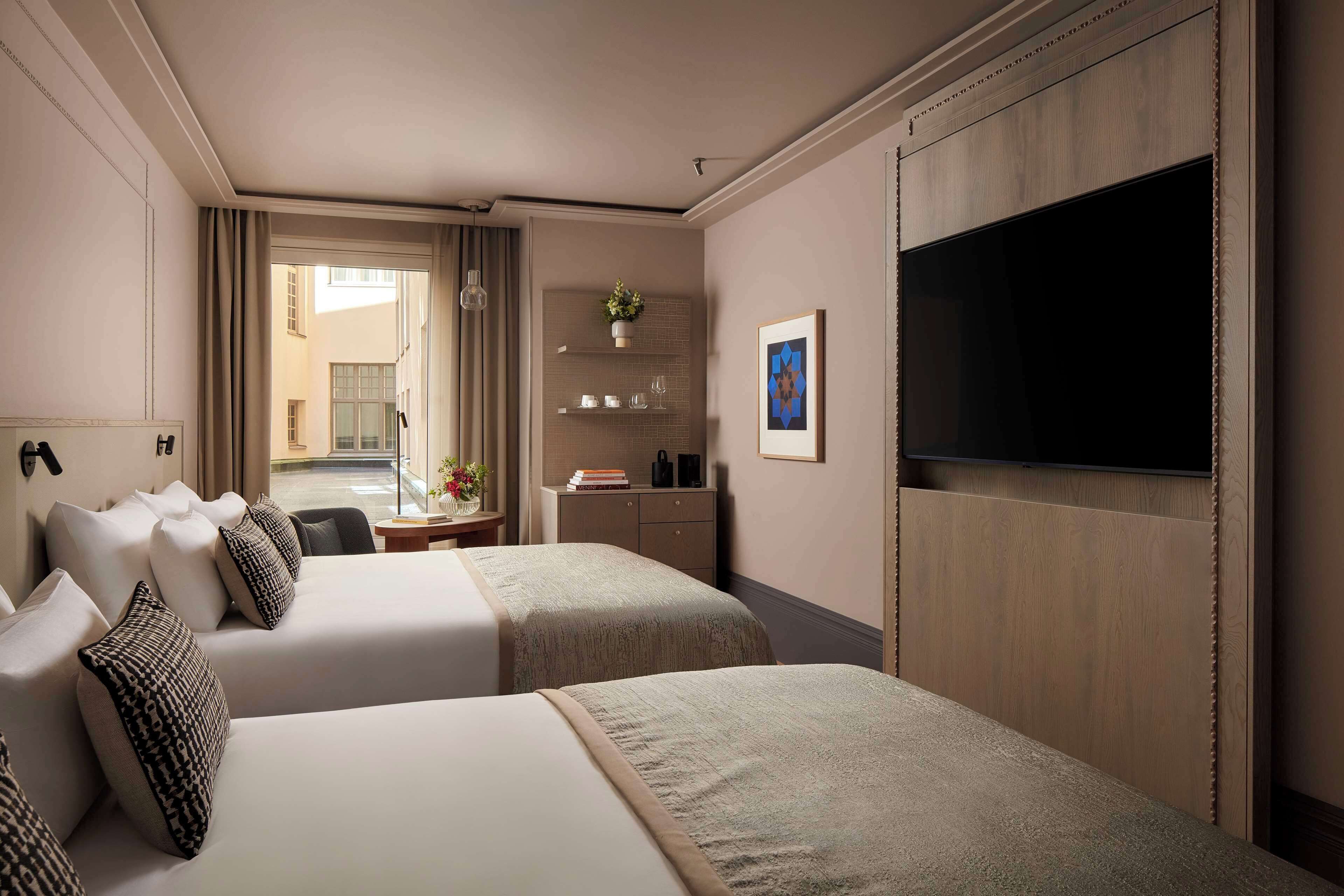 A hotel room with two beds, a TV, a desk with a chair, a small table with a plant, and a view of the outside.
