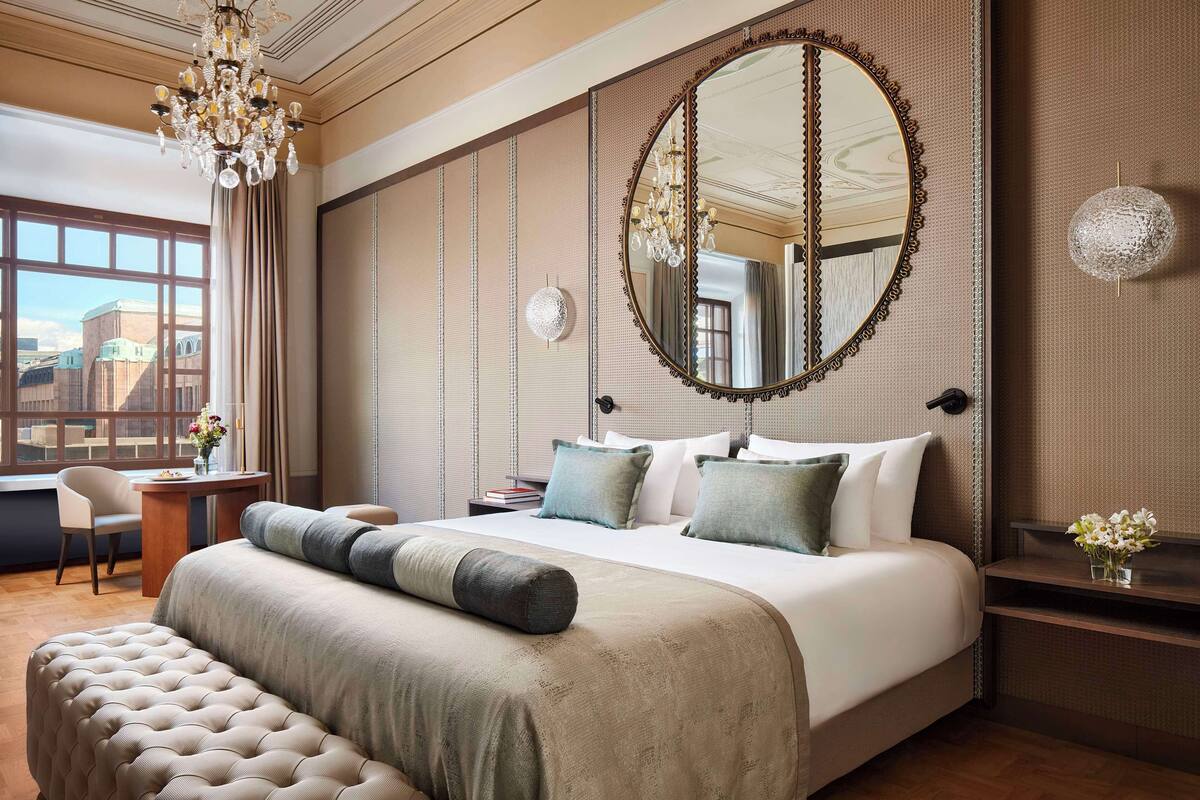Ein Hotelzimmer mit einem großen Bett, einer Kandelaber, einem runden Spiegel und Blick auf die Stadt durch große Fenster.