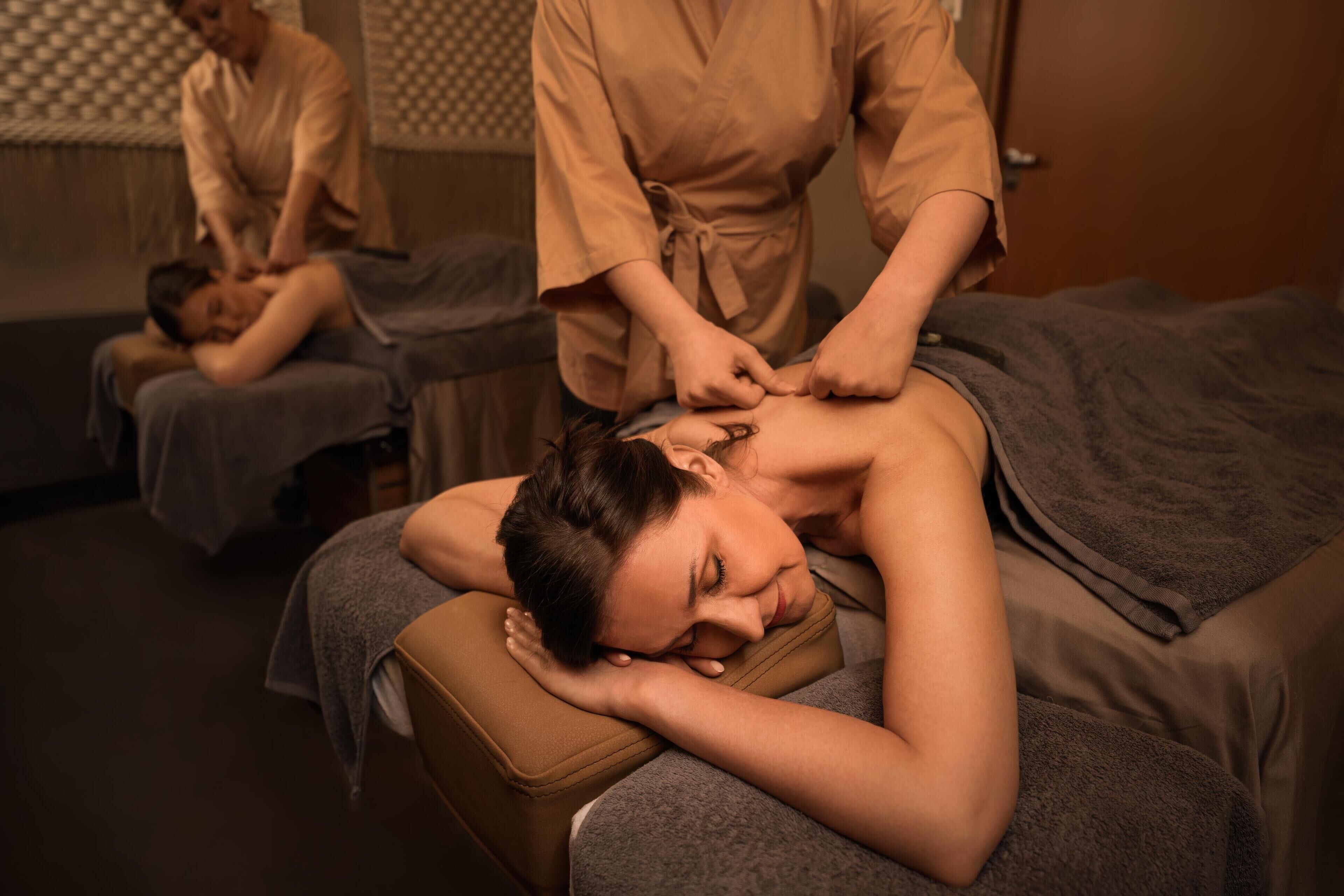 A massage therapy room with a client lying on a massage table, a therapist attending to the client, and another client in the background.