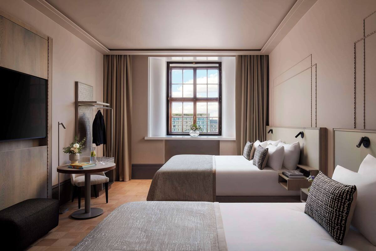 Ein Hotelzimmer mit einem großen Bett, einem Einzelbett, einem Schreibtisch mit Stuhl, einem Fernseher und einem Fenster mit Vorhängen.