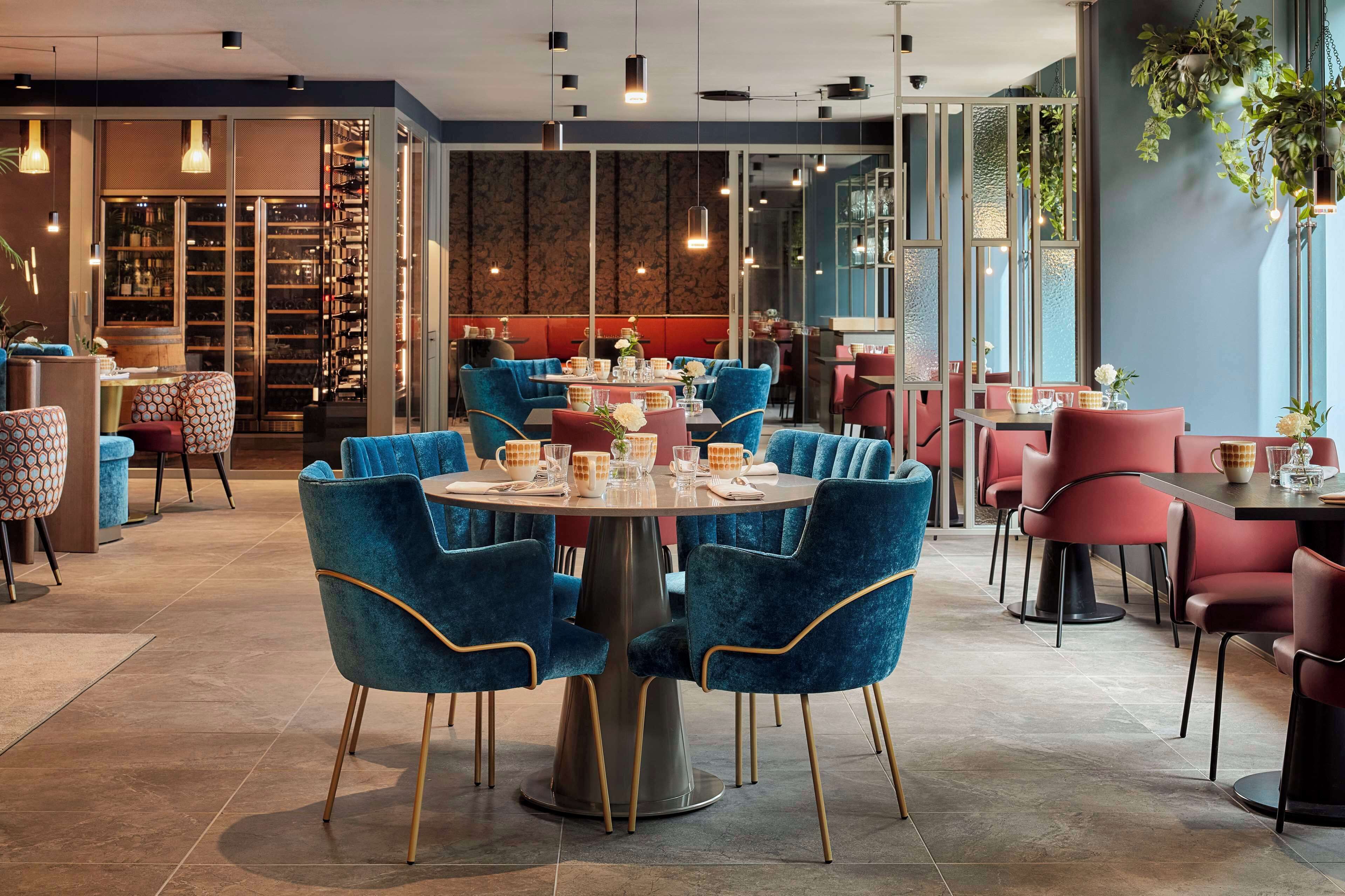 A modern restaurant with round tables, blue and red chairs, and a wine cellar visible through glass doors.