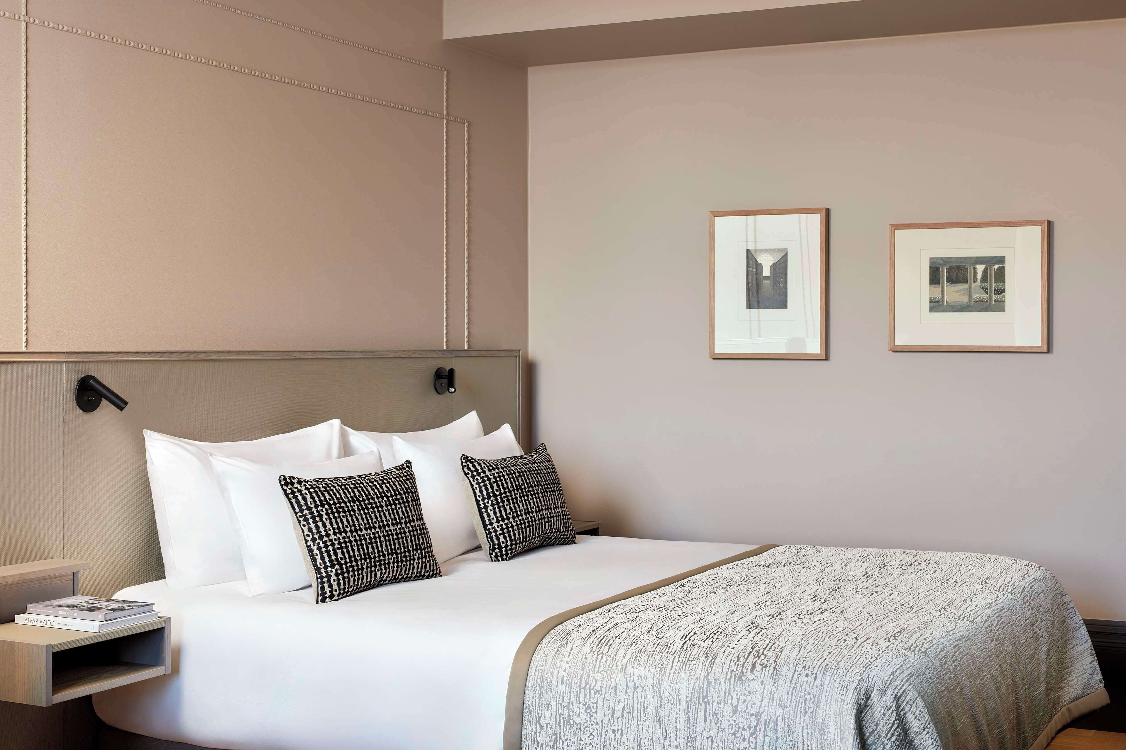 A neatly made bed with white linens and patterned pillows, a bedside table with a book, and two framed pictures on the wall.