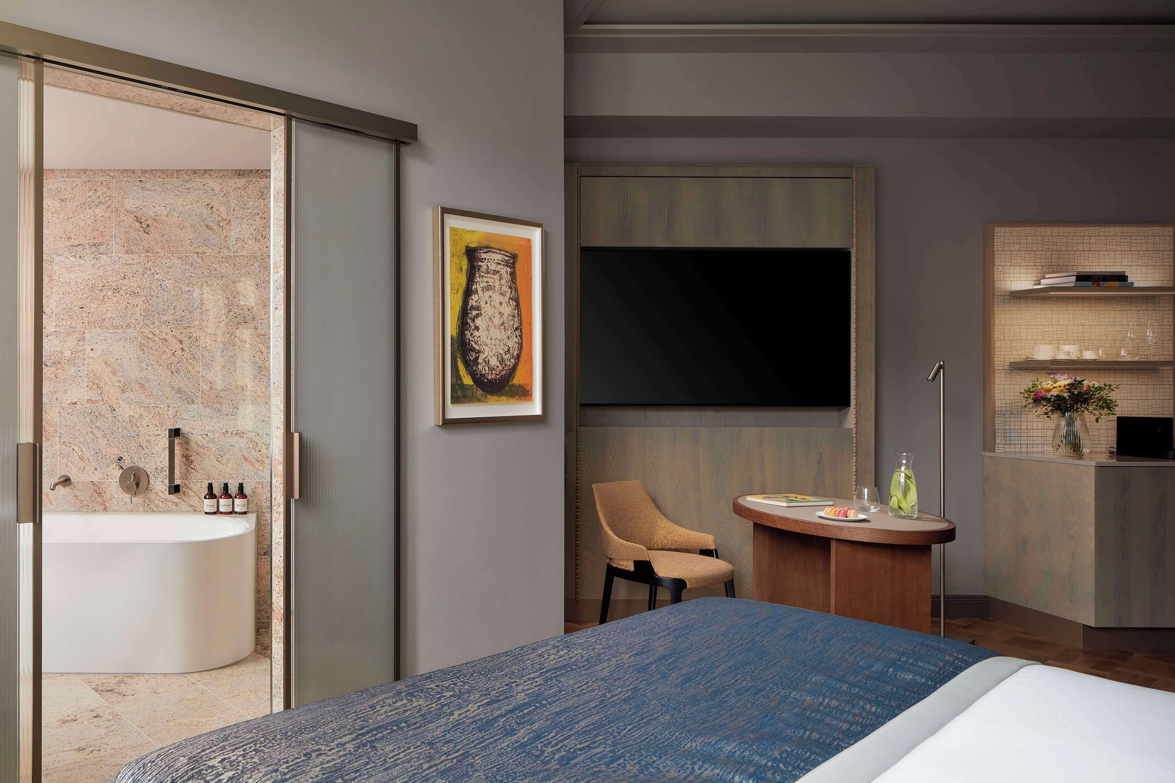 Una camera d'hotel moderna con un letto grande, un bagno con vasca, un tavolino rotondo con una sedia e uno scaffale con libri e oggetti decorativi.