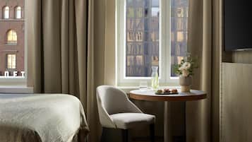 Una habitación de hotel con una cama, una silla, una mesita con un jarrón de flores y una ventana que da a un edificio de ladrillo.