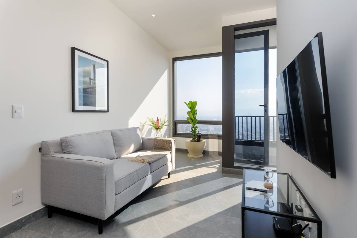 A modern living room with a grey sofa, a framed picture, a large TV, a glass coffee table, and a potted plant.