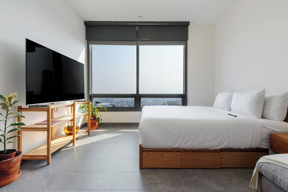 A modern bedroom with a large bed, a TV on a stand, a desk, and a window with a city view.