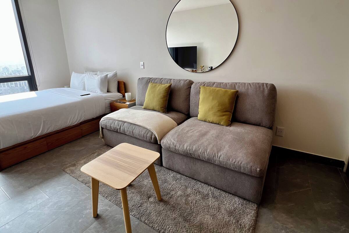 A modern hotel room with a bed, a sofa, a small table, and a round mirror on the wall.