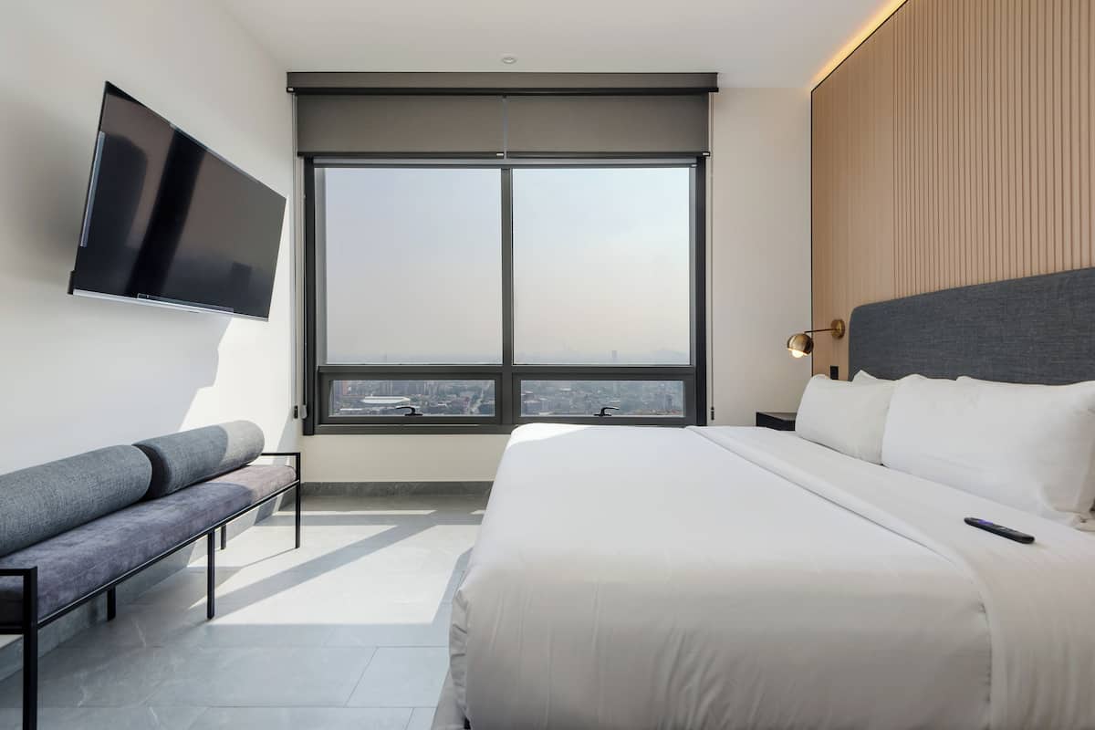 A modern hotel room with a large bed, a flat-screen TV mounted on the wall, a bench, and a large window offering a city view.