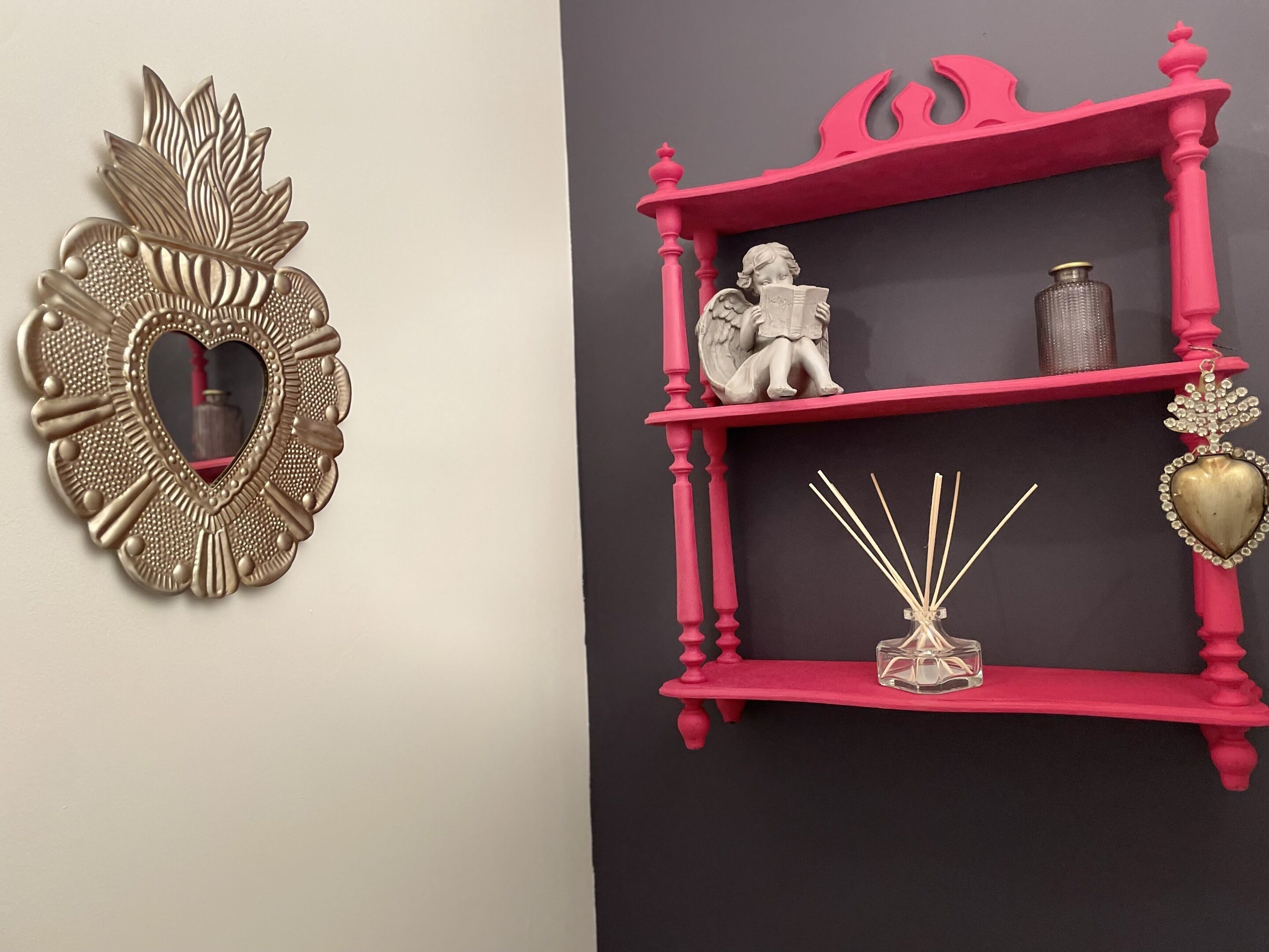 Una pared con un espejo decorativo con forma de corazón y un estante rosa con una estatua, un jarrón y un adorno con forma de corazón.