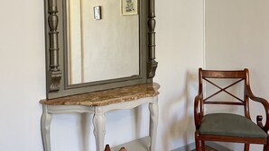 A mirror with a wooden frame and a marble top, mounted on a white console table with a shelf below. A wooden chair with a cushioned seat is positioned to the right of the mirror.